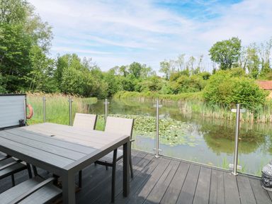 A patio with a table and chairs overlooking a pond at Dragonfly Lodge in Whinburgh near Dereham