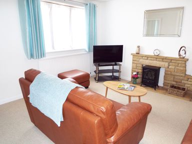 A living room with a brown leather sofa, light blue curtains, a TV on a stand, a wooden coffee table with magazines, and a stone fireplace with a small stove at Kates Cottage in Bourton-on-the-Water