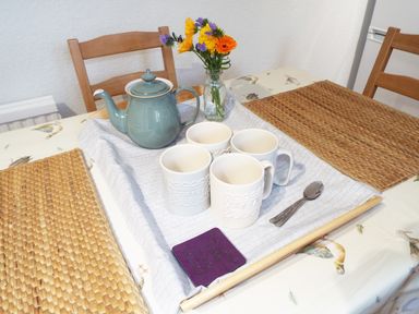 A kitchen table with a green teapot four white mugs a small vase with flowers and two spoons at Kates Cottage in Bourton-on-the-Water