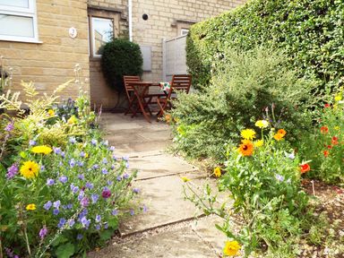 An outdoor patio with wooden table and chairs surrounded by flowering plants and greenery at Kates Cottage in Bourton-on-the-Water