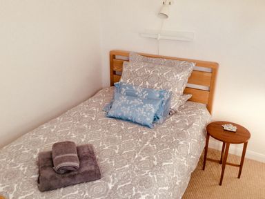 A bedroom with a single bed with patterned bedding and two blue decorative pillows with a wooden headboard and a small round wooden side table with coasters in Kate's Cottage in Bourton-on-the-Water