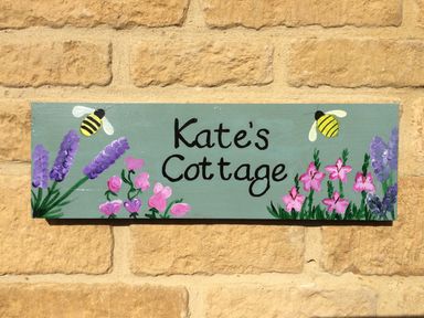 A painted sign with flowers and bees on a brick wall at Kates Cottage in Bourton-on-the-Water