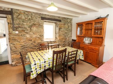 A dining room with a wooden table covered with a polka dot tablecloth and wooden chairs next to a stone wall with a window and a wooden cabinet at Nantmel in Beddgelert