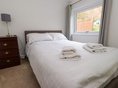 A bedroom with a double bed white bedding folded towels on bed wooden nightstand with lamp beside window with curtains at Nantmel in Beddgelert
