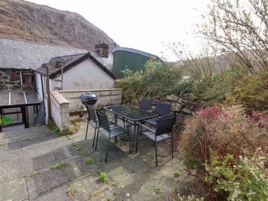An outdoor patio with a glass table and six chairs a barbecue grill stone wall and bushes near houses with a mountain in the background at Nantmel in Beddgelert