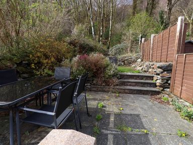 An outdoor patio with a glass table and chairs surrounded by trees and bushes at Nantmel in Beddgelert