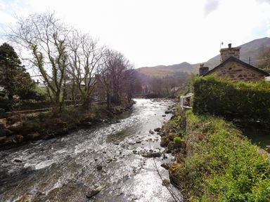 A river flowing between trees and houses with hills in the background at Nantmel in Beddgelert