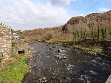 A river with rocks flowing beside stone buildings and trees with hills in the background at Nantmel in Beddgelert