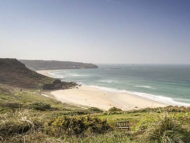 A beach with waves and cliffs in the background with grass and a wooden bench in the foreground at Tregiffian Vean in Sennen Cove