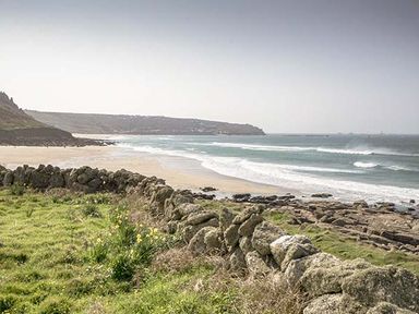 A coastal landscape with a stone wall in the foreground a sandy beach waves and cliffs at Tregiffian Vean in Sennen Cove
