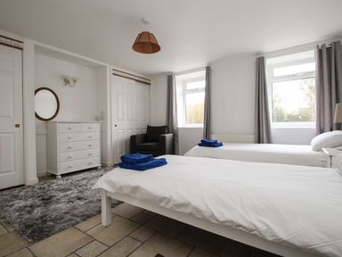 A bedroom with two single beds a white dresser a gray armchair and two windows with curtains at Tregiffian Vean in Sennen Cove