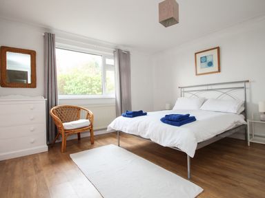 A bedroom with a double bed with white bedding and blue towels a wooden chair white chest of drawers and a window with grey curtains at Tregiffian Vean in Sennen Cove