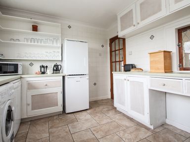 A kitchen with white cabinets a white fridge a microwave glass shelves and a wooden bread box at Tregiffian Vean in Sennen Cove