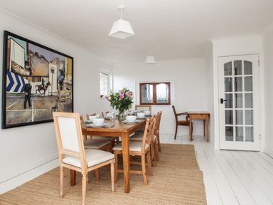 A dining room with a wooden table set with white dishes and a vase of flowers a large painting on the wall and a small wooden desk at Tregiffian Vean in Sennen Cove