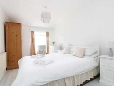 A bedroom with a white bed holding pillows and towels two white bedside tables with lamps a wooden wardrobe a white dresser with flowers and a window with curtains at The Coach House in Sennen near St Just