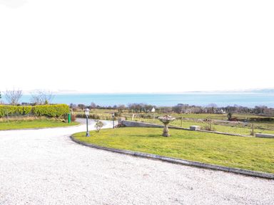 A gravel driveway curving around a lawn with a birdbath and lamp posts overlooking fields and a body of water at Millers Lane House in Muff County Donegal