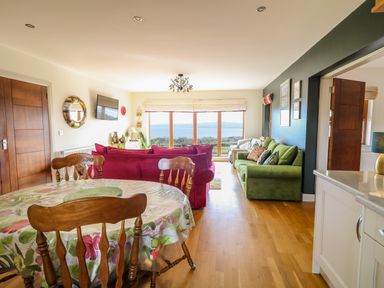 A living room and dining area with wooden chairs a floral tablecloth on the table red and green sofas a wall-mounted television and large windows overlooking a scenic view at Millers Lane House in Muff County Donegal
