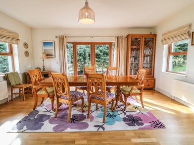 A dining room with wooden table and six chairs on a floral rug next to windows and glass cabinets at Millers Lane House in Muff County Donegal