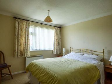 A bedroom with a bed with a metal headboard bedside tables with lamps a window with curtains and a chair in Skylark Weybourne