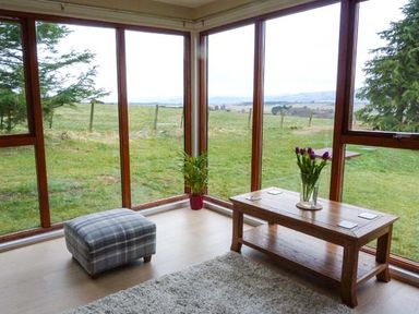 A corner room with large windows a wooden coffee table with flowers a plaid ottoman and a plant at Pine Lodge in Culbokie