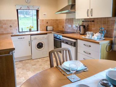 A kitchen with a washing machine oven and dishwasher and a dining table with plates and a glass at Pine Lodge in Culbokie