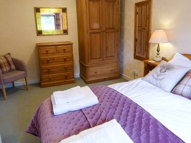 A bedroom with a made bed and folded towels on it a wooden wardrobe chest of drawers a purple armchair and a lamp at Pine Lodge in Culbokie