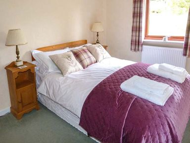 A bedroom with a double bed white bedding purple throw pillows wooden side tables with lamps and a window with plaid curtains at Pine Lodge Culbokie