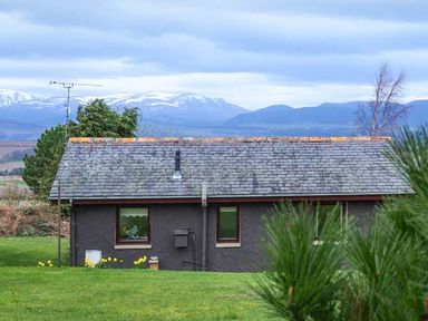 A small house with a slate roof surrounded by grass with mountains in the background at Pine Lodge in Culbokie