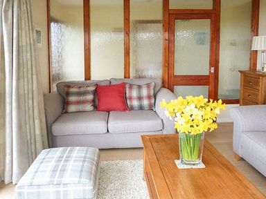 A living room with a sofa with cushions a wooden coffee table with a vase of yellow flowers a plaid ottoman and a wooden cabinet with a lamp at Pine Lodge in Culbokie
