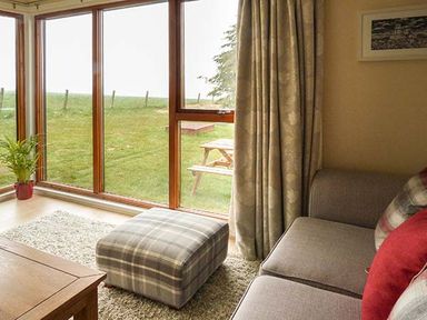 A living room with a sofa a plaid ottoman a wooden table and large windows overlooking a grassy yard at Pine Lodge in Culbokie
