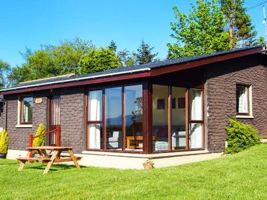A single-story house with large windows and a wooden picnic table on a lawn at Pine Lodge in Culbokie