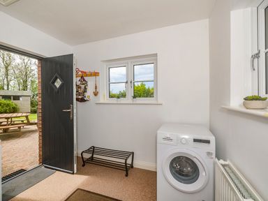 A utility room with a washing machine a shoe rack a coat rack an open black door and two windows at Penbarden Barn in Dizzard near Crackington Haven