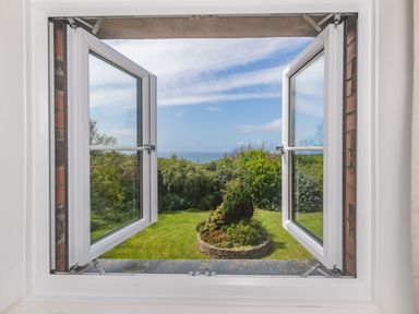 View through open white framed window onto garden with circular brick flower bed and trees at Penbarden Barn in Dizzard near Crackington Haven
