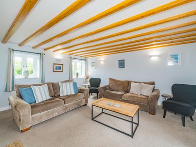 A living room with two brown sofas, two black chairs, a wooden coffee table, and windows with curtains at Penbarden Barn in Dizzard near Crackington Haven