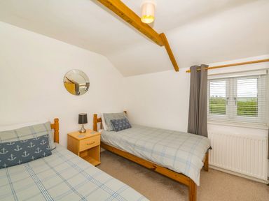 A bedroom with two single beds a nightstand with a lamp a window with blinds and curtains and a round mirror on the wall at Penbarden Barn in Dizzard near Crackington Haven