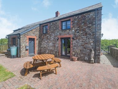 A stone house with brick accents and a wooden picnic table on a paved patio at Penbarden Barn in Dizzard near Crackington Haven