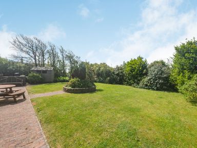 A garden with a grass lawn a circular stone planter trees shrubs a wooden shed bench and picnic table at Penbarden Barn in Dizzard near Crackington Haven