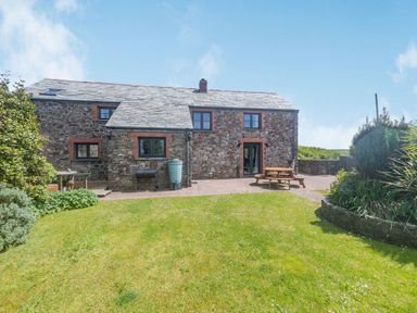 A stone house with a slate roof and garden furniture on a patio with a lawn and plants at Penbarden Barn in Dizzard near Crackington Haven