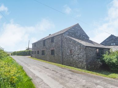 A stone barn building next to a country road with grass and shrubs at Penbarden Barn in Dizzard near Crackington Haven