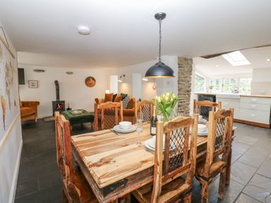 A dining area with a wooden table set for six with a vase of flowers and a bottle with an open plan kitchen and living room featuring sofas and a wood burning stove at The Dale Cottage in Allendale near Hexham