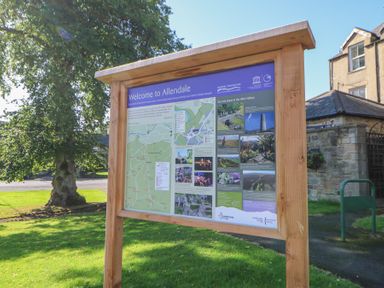 A wooden information board with maps and text on a grassy area near trees and stone buildings at The Dale Cottage in Allendale near Hexham