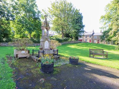 An outdoor park area with a stone monument surrounded by flowers wooden planters a bench and houses in the background at The Dale Cottage in Allendale near Hexham