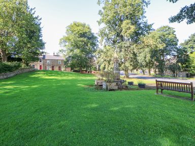 A grassy park area with a stone monument planter and a wooden bench near houses and trees at The Dale Cottage in Allendale near Hexham