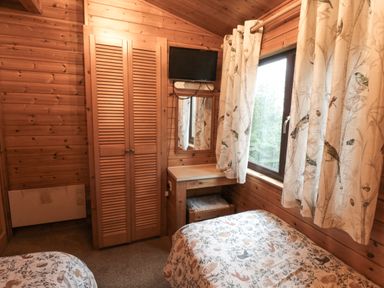 A wooden bedroom with two beds a wardrobe a window with patterned curtains a small desk with a mirror and a mounted television at Sycamore Lodge in Liverton near Danby