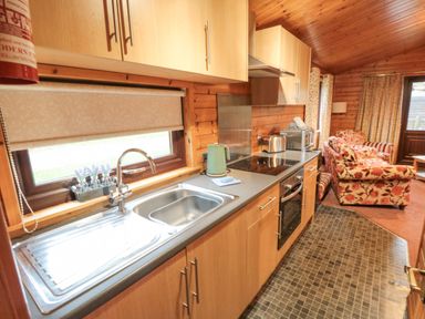 A kitchen with wooden cabinets a stainless steel sink a stovetop and oven a kettle toaster and microwave next to a living area with floral patterned sofas at Sycamore Lodge in Liverton near Danby