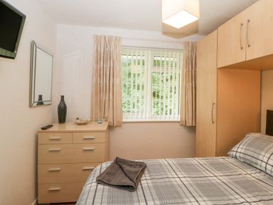 A bedroom with a bed with checked bedspread a chest of drawers a mirror curtain covered window and built in wardrobe at Sycamores Rosecraddoc Lodge near Liskeard