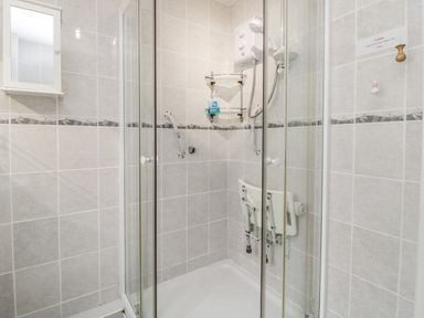 A tiled shower enclosure with a folding seat a wall-mounted shower unit and grab bars at Sycamores Rosecraddoc Lodge near Liskeard