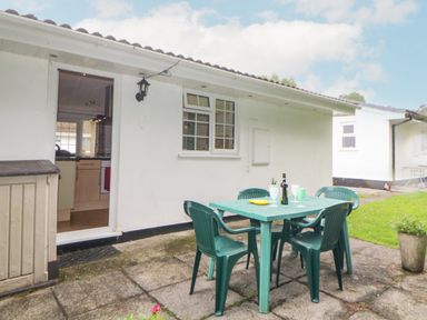 An outdoor patio with a green plastic table and four chairs near a white building with an open door and window at Sycamores in Rosecraddoc Lodge near Liskeard