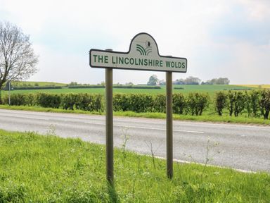 A road sign for The Lincolnshire Wolds beside a road with green fields and hedges in the background