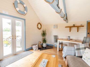A living room with a wooden floor a wooden table two plaid chairs a sofa with pillows indoor plants a wicker dog three wall decorations and glass doors at The Nook in Louth